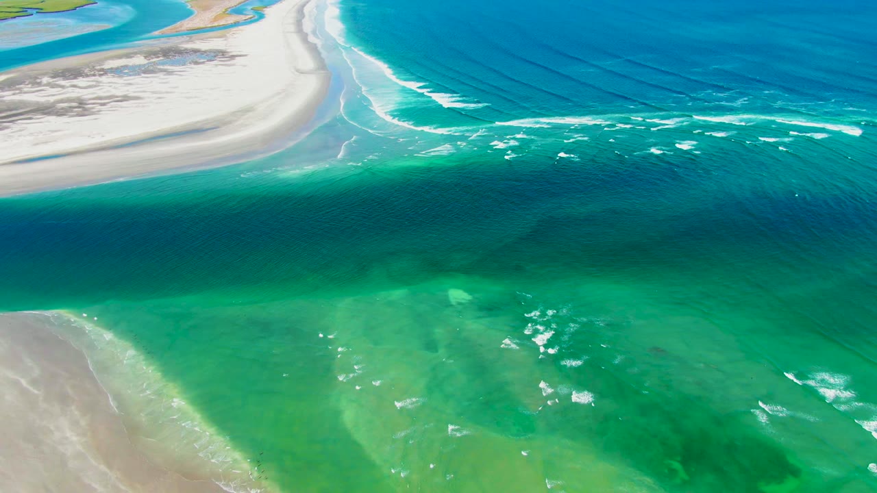 Aerial frame over La Bocana, Baja California Sur, showing pale sand at the upper left, turquoise shallows below, darker blue water offshore, and no visible people.