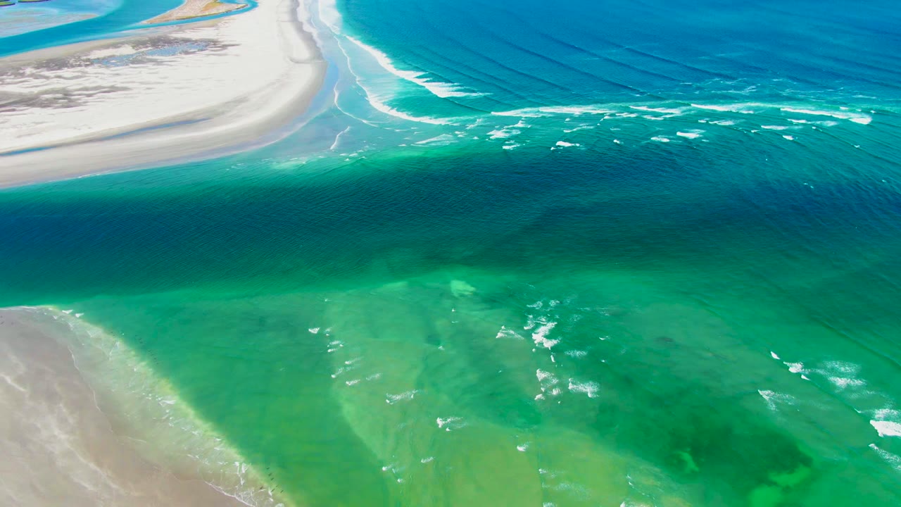 Aerial frame over La Bocana, Baja California Sur, with sandy shoreline near the top left, diagonal surf lines in blue water, green shallows in the foreground, and no visible people.
