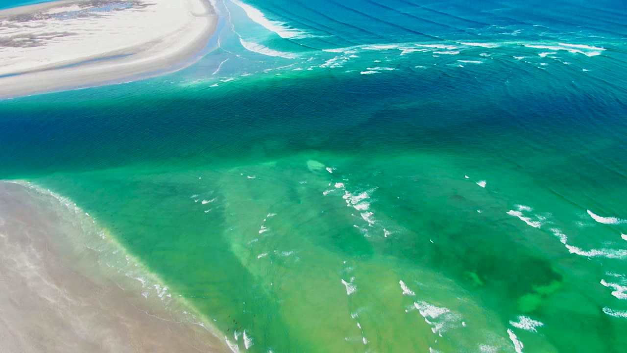 Aerial frame from La Bocana, Baja California Sur showing turquoise shallow water between pale sandbars, small white foam streaks, deeper blue water above, and no visible people.