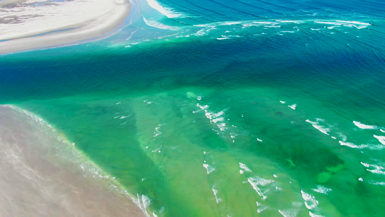Aerial frame from La Bocana, Baja California Sur showing turquoise shoreline water, pale sand along the left and upper edges, scattered white foam streaks, darker blue sea beyond, and no visible people.