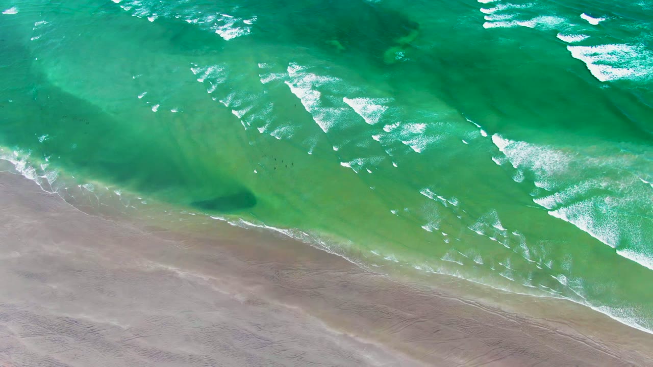 Aerial view over La Bocana showing turquoise ocean foam sweeping diagonally toward wet sand, with tiny dark birds near the shoreline and no visible people.