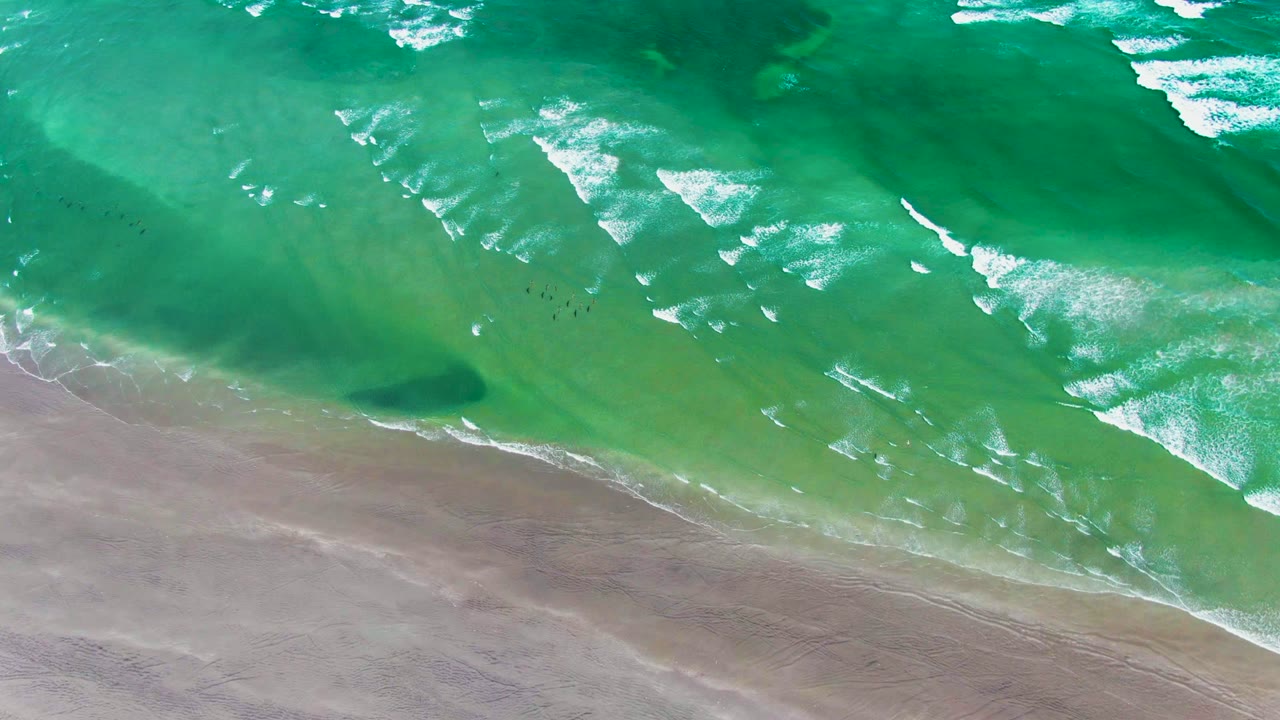 White surf lines shift across green water beside a sandy shore.