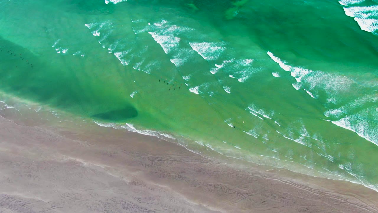 Overhead La Bocana shoreline view with bright foam patches scattered across turquoise water and smooth wet sand occupying the lower part of the frame, with no visible people.