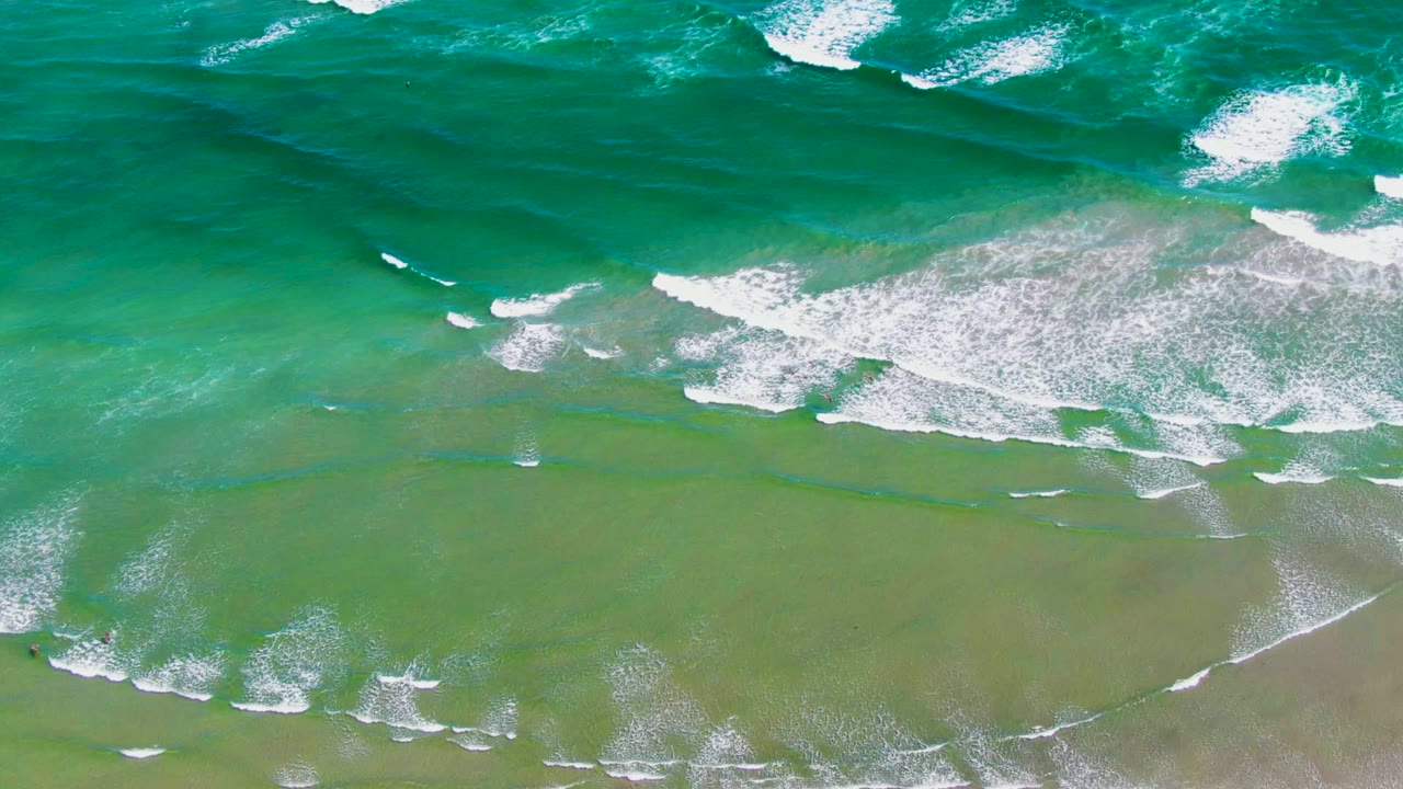 La Bocana surf seen from above, with white breakers spreading across the middle of the frame, green water near the beach, turquoise water offshore, and no clearly visible people.