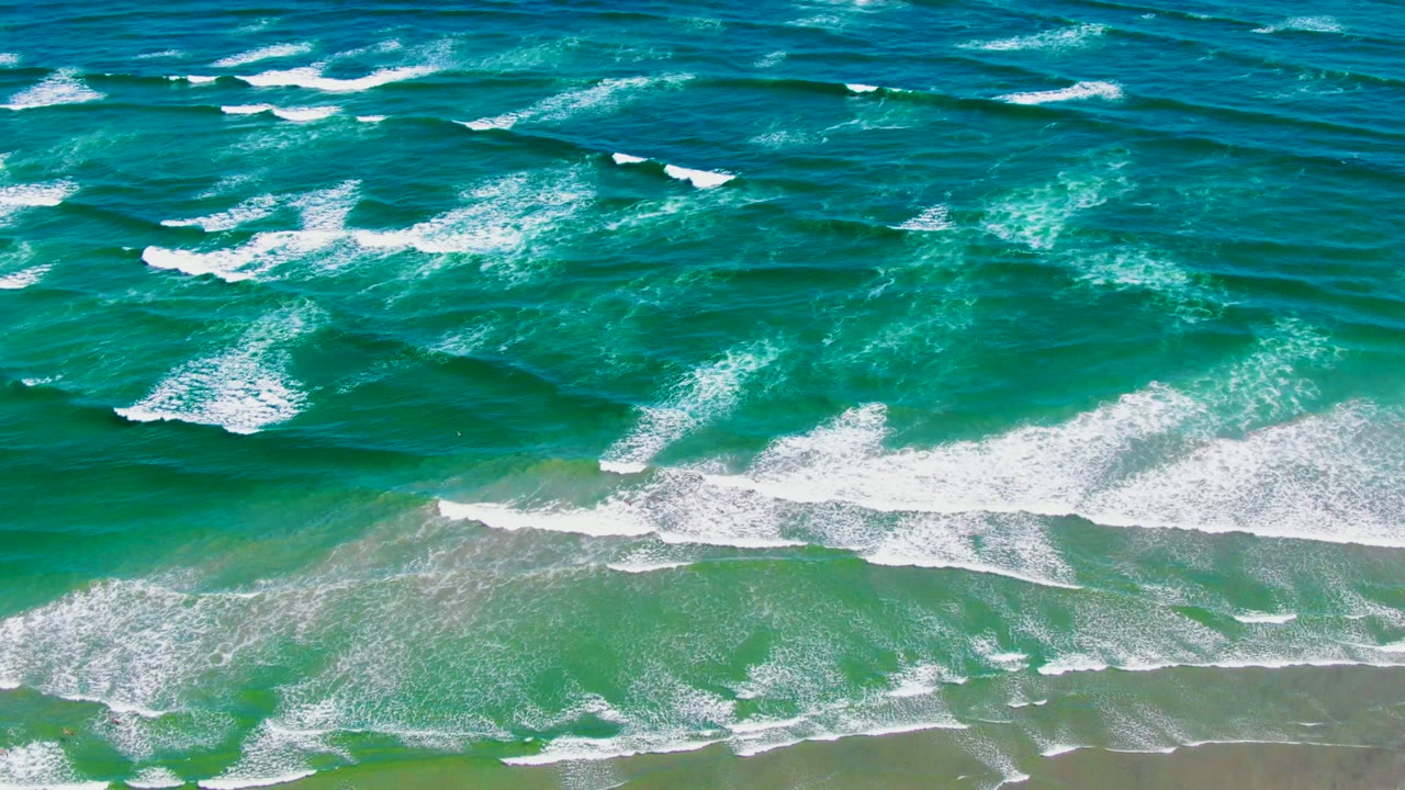 At 0:00 in La Bocana, Baja California Sur, deep blue ocean shifts to turquoise surf as white foam bands approach wet sand, with no visible people.