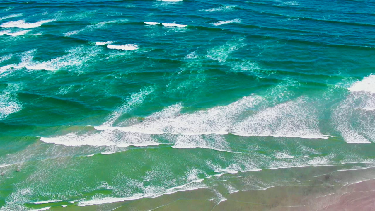 At 0:02 in La Bocana, Baja California Sur, diagonal rows of white foam cross the turquoise water and thin out over the wet sand, with no visible people.
