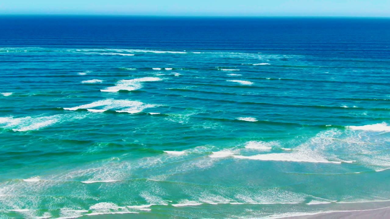 At La Bocana, Baja California Sur, deep blue water meets turquoise surf bands rolling toward a pale sandy shore, with no visible people.
