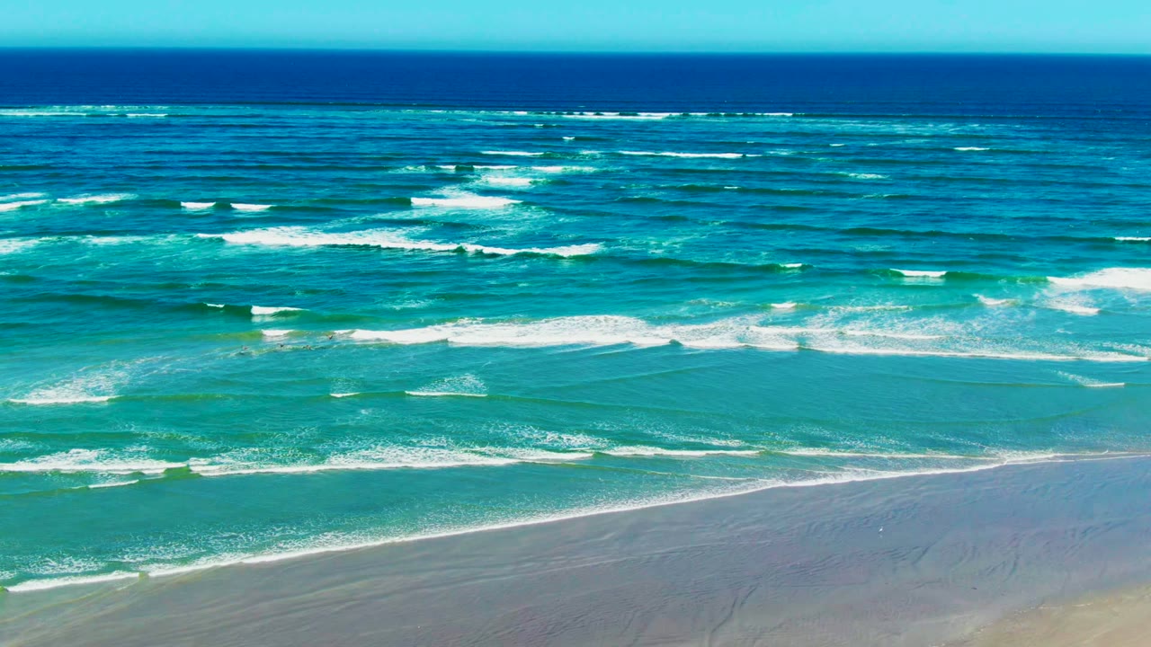 Wide elevated view of La Bocana ocean surf, with deep blue water near the horizon, turquoise shallows, white foam bands, sandy beach at the bottom, and no visible people.