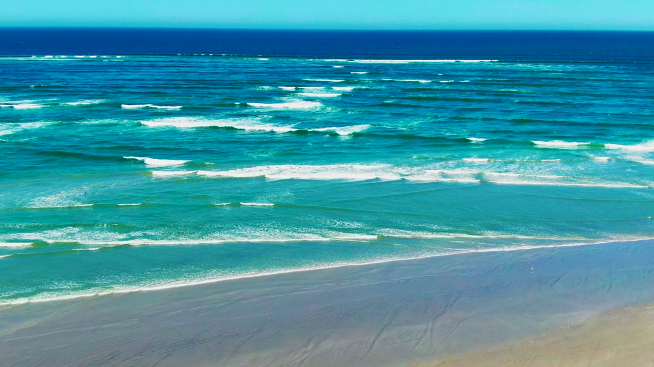 At La Bocana, white foam bands spread across blue-green water, with the sandy foreground partly washed by shallow surf and no visible people or structures.