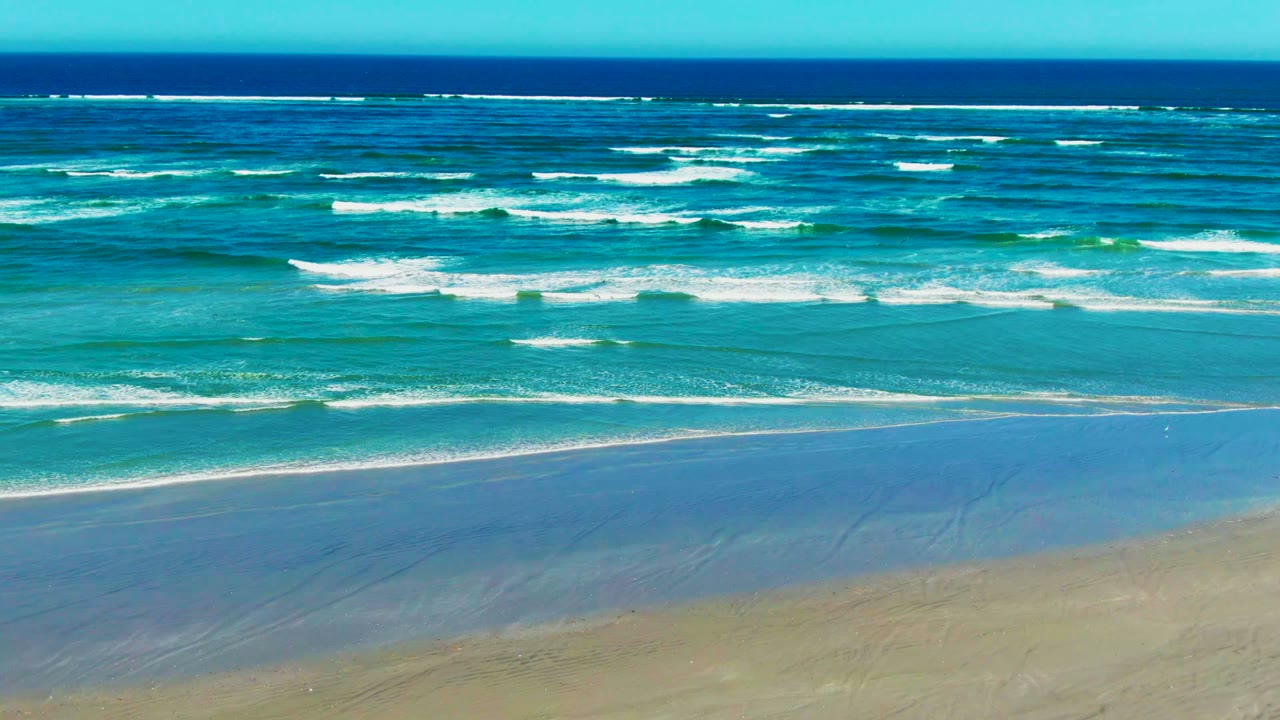 Wide view of La Bocana beach with tan wet sand, turquoise water bands, white breaking waves, deep blue horizon, and no visible people.