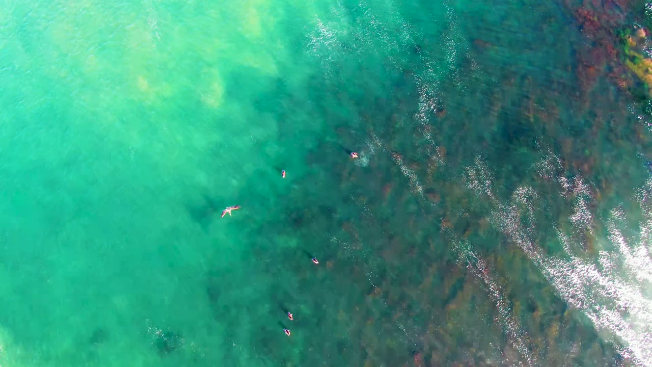 Overhead view at La Bocana, Baja California Sur showing several small swimmers spread across clear turquoise water with darker seabed bands and bright surface reflection.