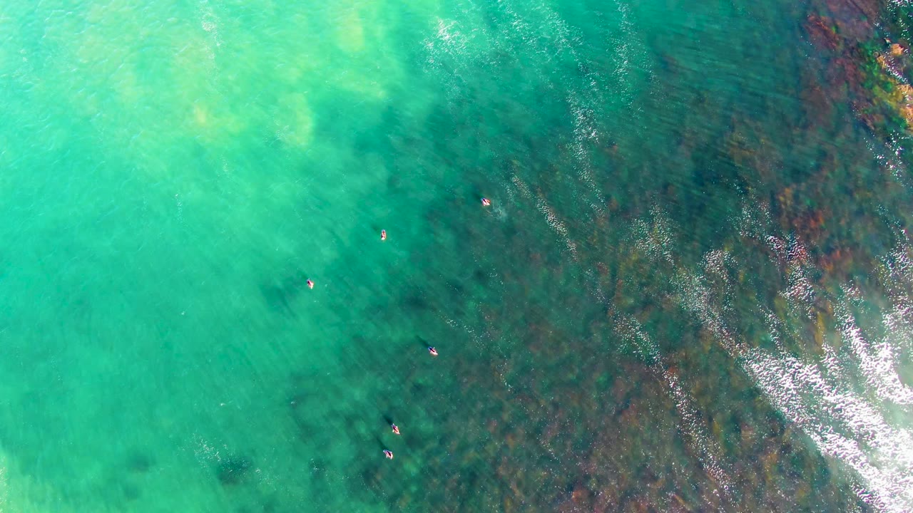 Top-down La Bocana water view showing tiny swimmers against turquoise shallows, with darker underwater texture and a bright ripple line near the right edge.