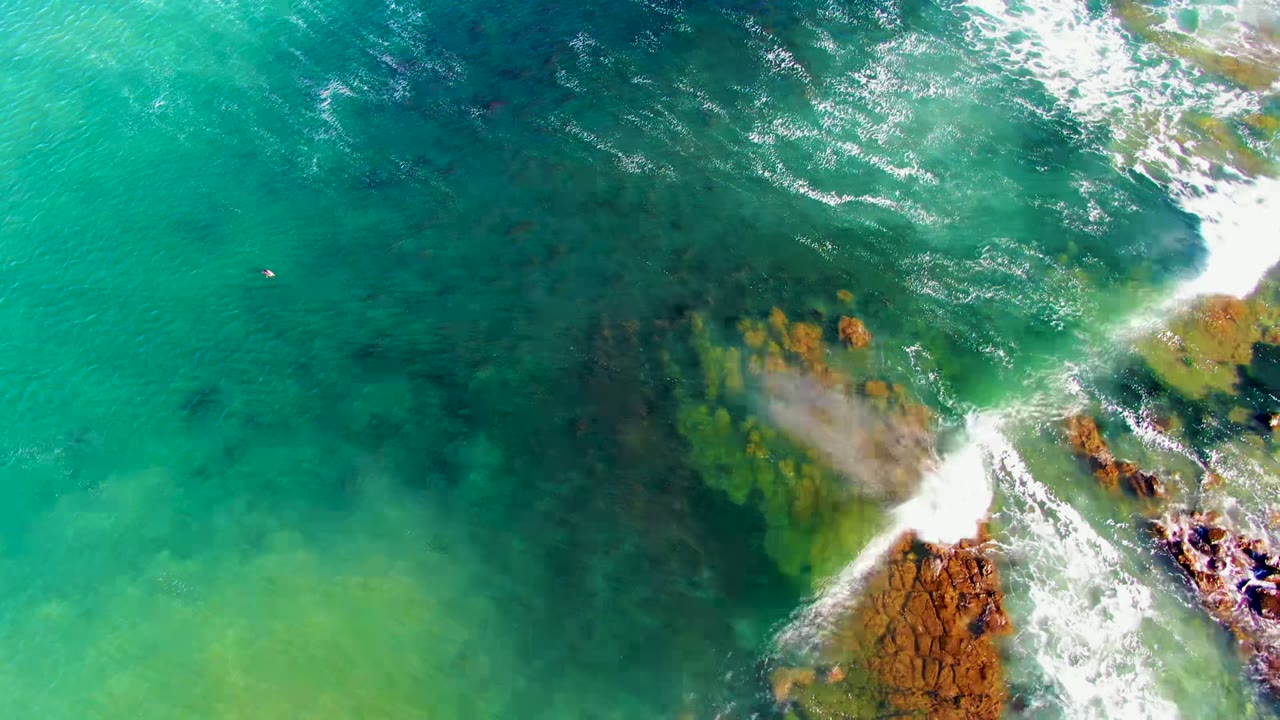 Overhead view in La Bocana, Baja California Sur of turquoise water, submerged dark patches, and white surf breaking around rust-colored rocks, with no visible people.