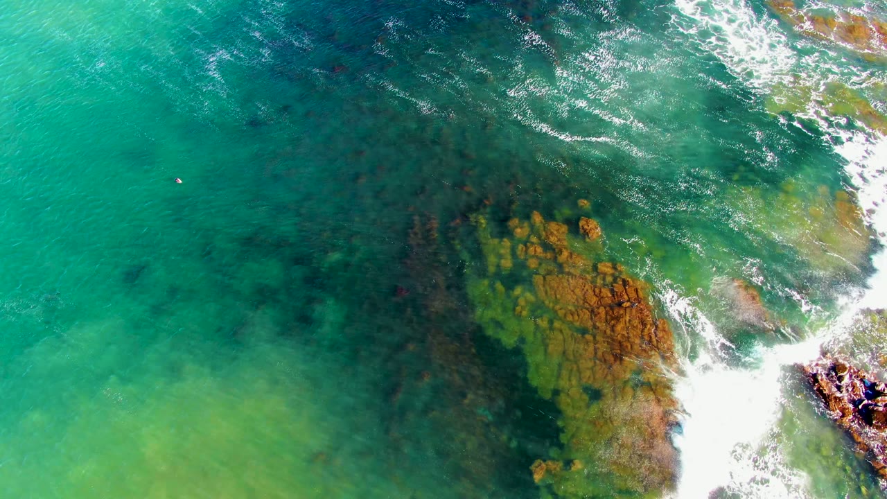 High overhead view in La Bocana of white wave wash crossing orange-brown rocks beneath clear green water, with darker submerged patches to the left and no visible people.