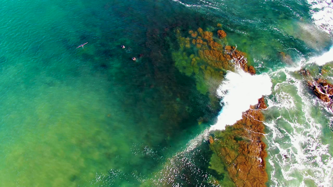 At La Bocana, the overhead frame shows a bird crossing open turquoise water while small swimmers remain near darker underwater rock and white surf on the right.