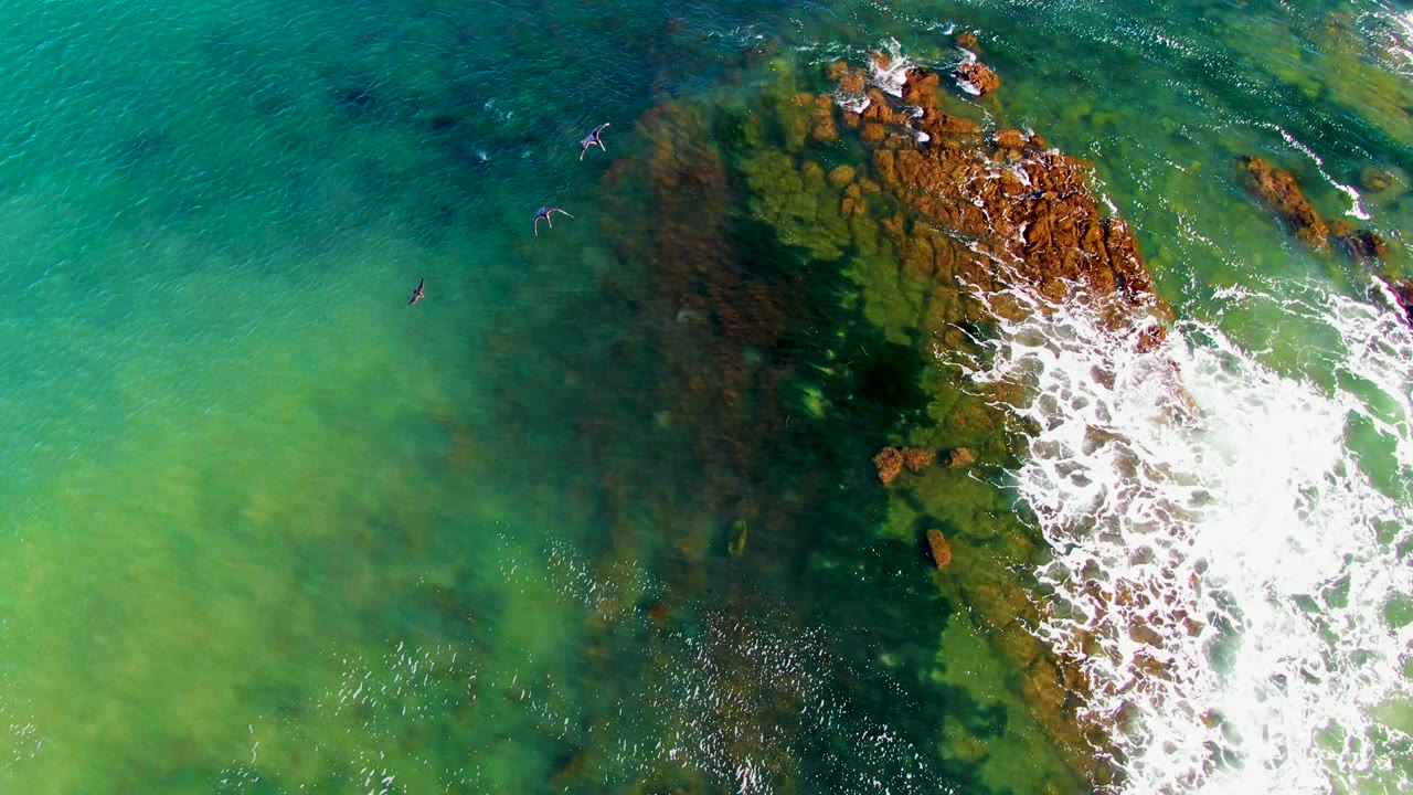 Overhead frame from La Bocana, Baja California Sur showing small swimmers in clear green and turquoise water beside brown rocks with white surf on the right.