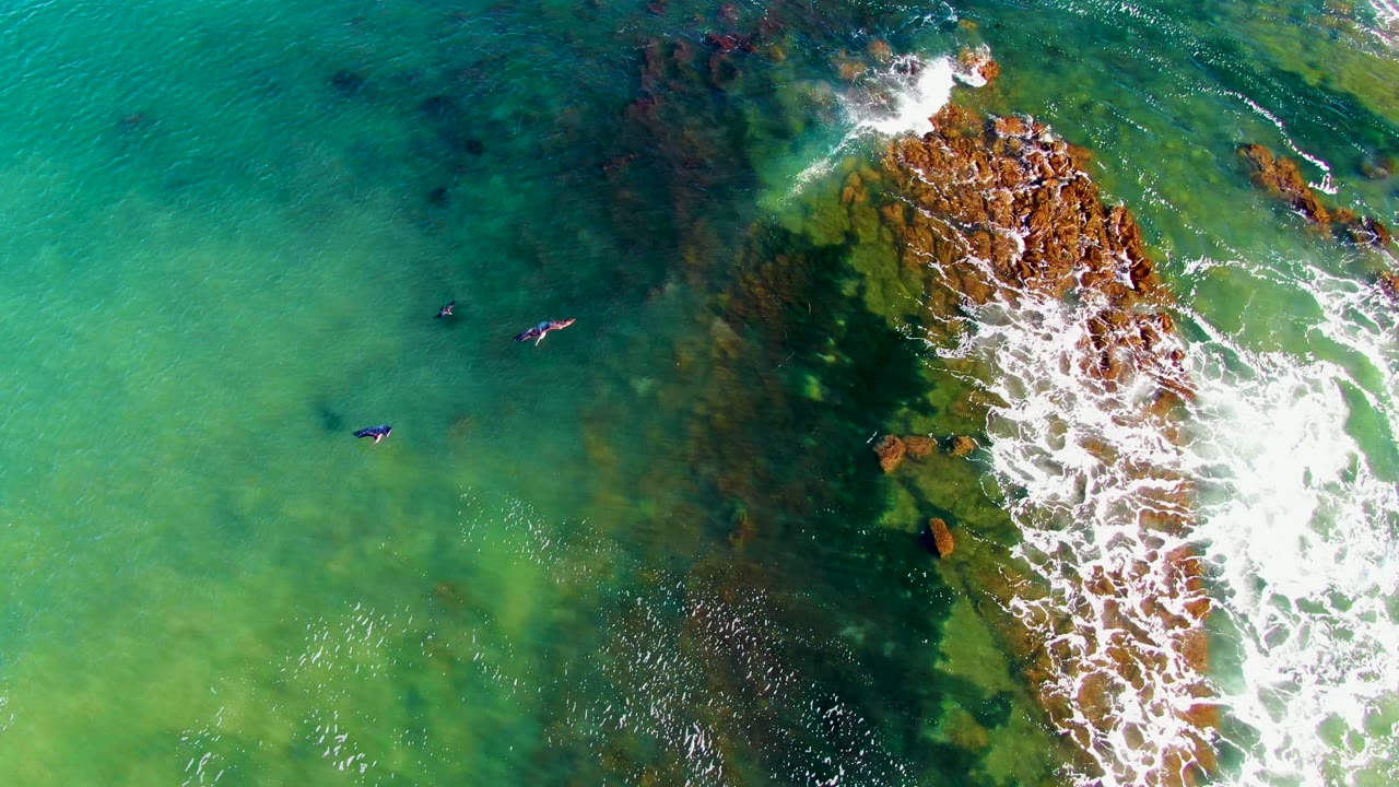 La Bocana aerial frame with two small swimmers near center-left, darker underwater patches below them, and a foamy rock line along the right edge.
