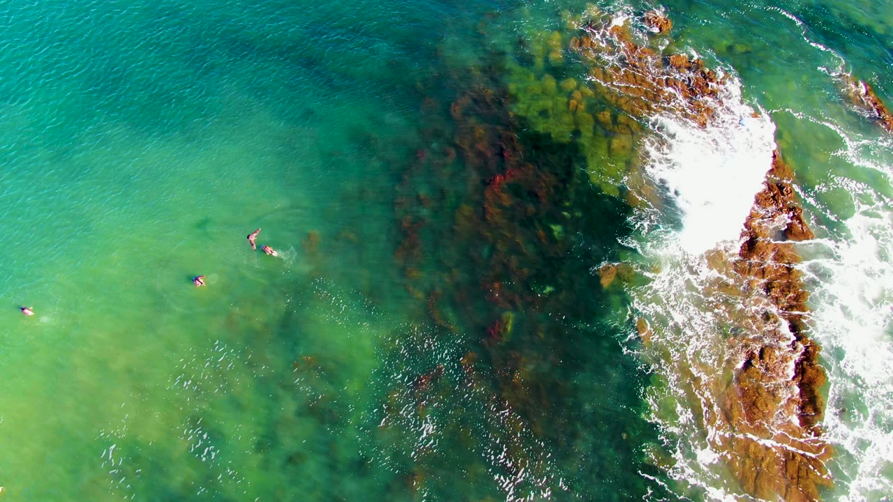 Overhead view in La Bocana, Baja California Sur showing small swimmers in turquoise water left of a rocky surf break, with white foam spreading across the rocks.