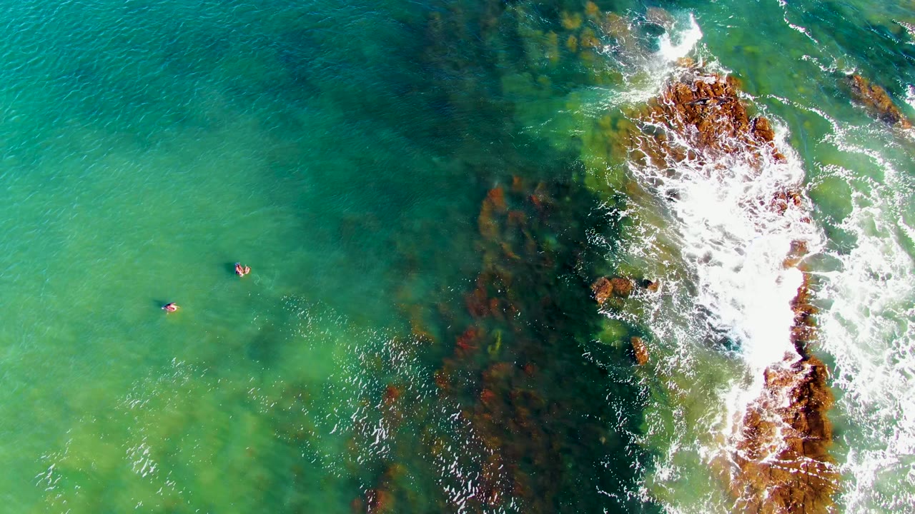 Overhead La Bocana view with swimmers near the center-left, dark underwater rock shadows through clear water, and white water curling around the rocks on the right.