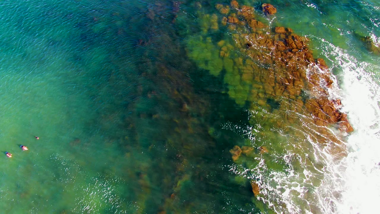 Overhead view of clear turquoise water in La Bocana, Mexico, with dark submerged rocks, white foam on the right, and tiny distant swimmers near the left edge.