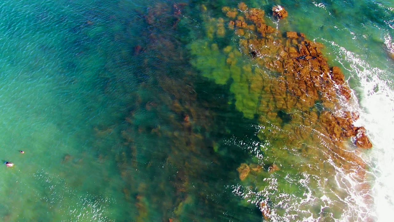 Aerial turquoise water over dark seabed with small swimmers near the lower left.