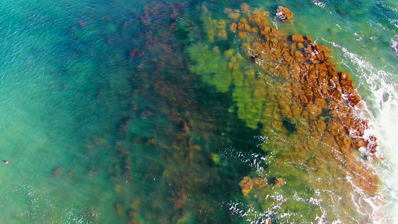 Top-down view in La Bocana with clear green-blue coastal water, dark underwater rock patterns, orange-brown rocks at the right, white foam, and no visible people.