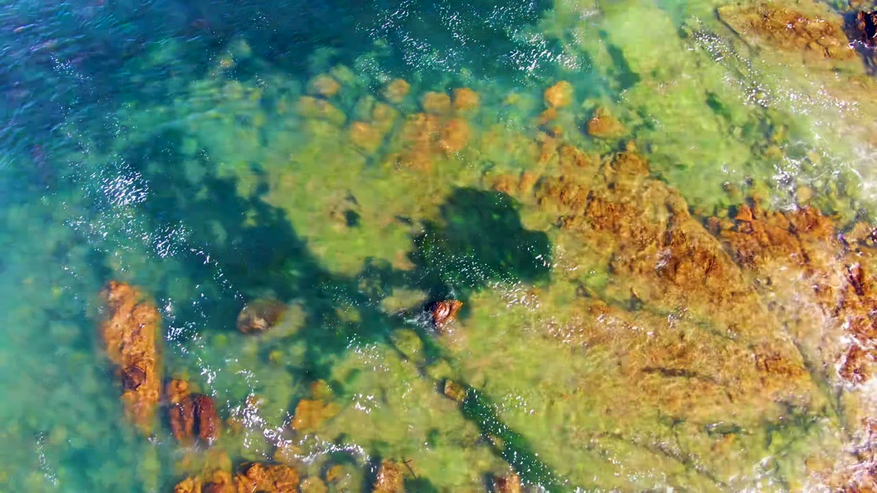 Top-down view of clear green-blue water over orange-brown rocks in La Bocana, Mexico, with a dark seabed patch near center and no visible people.