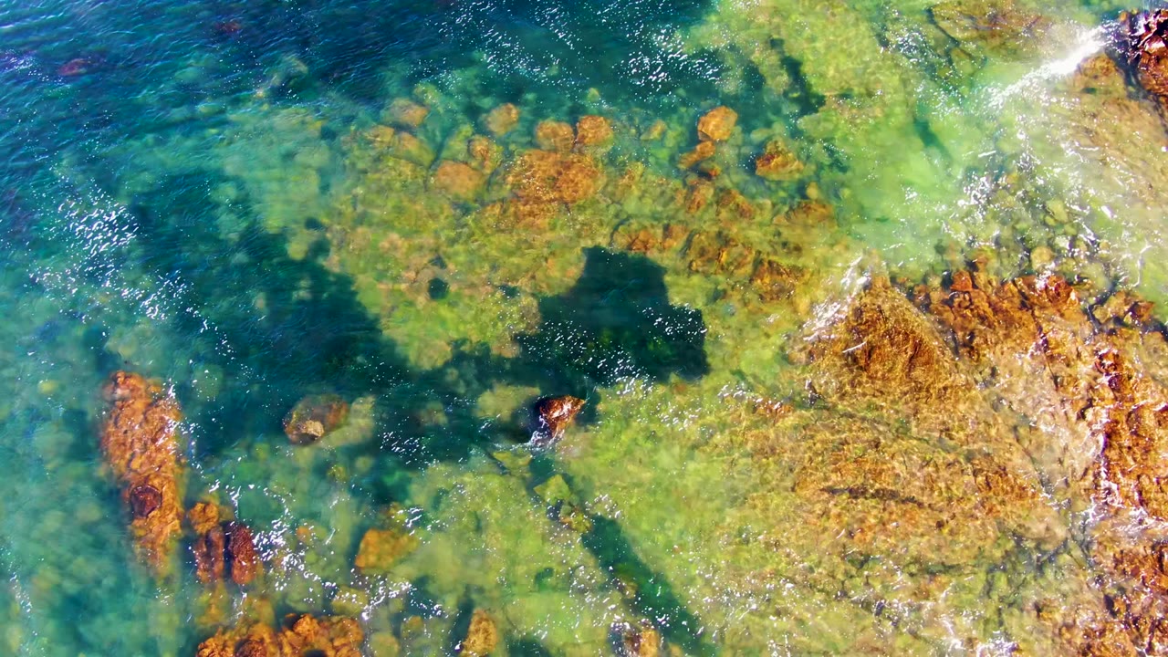 Top-down aerial frame in La Bocana, Mexico, with blue-green water, orange submerged rocks, and white foam beginning to enter along the right edge, with no visible people.