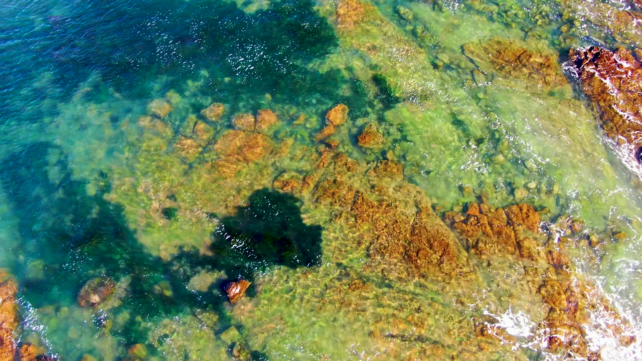 Top-down view of clear green and blue seawater over orange-brown rocks at La Bocana, with dark underwater patches and no visible people.