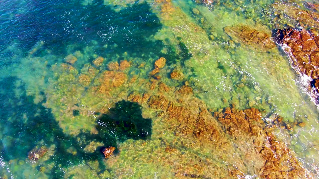 Aerial-looking view of green-blue shallow water and orange rock shelves at La Bocana, with white wash along the right edge and no visible people.