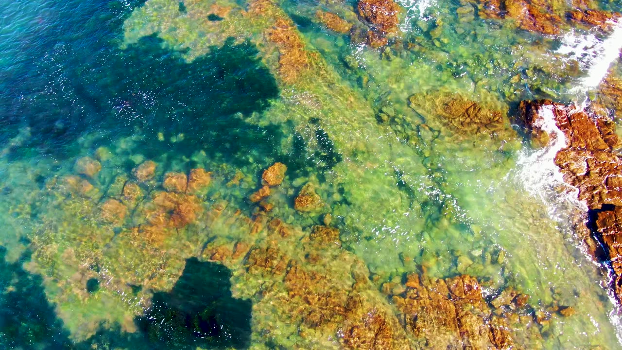 Aerial view in La Bocana, Baja California Sur, showing clear green-blue water over brown rocks, a darker patch of sea, and no visible people.