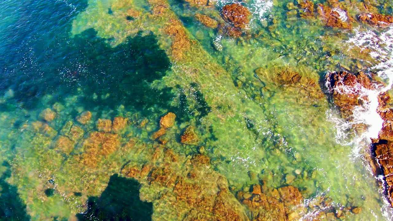 Aerial view of turquoise shallows with submerged rocks and foam along the rocks.