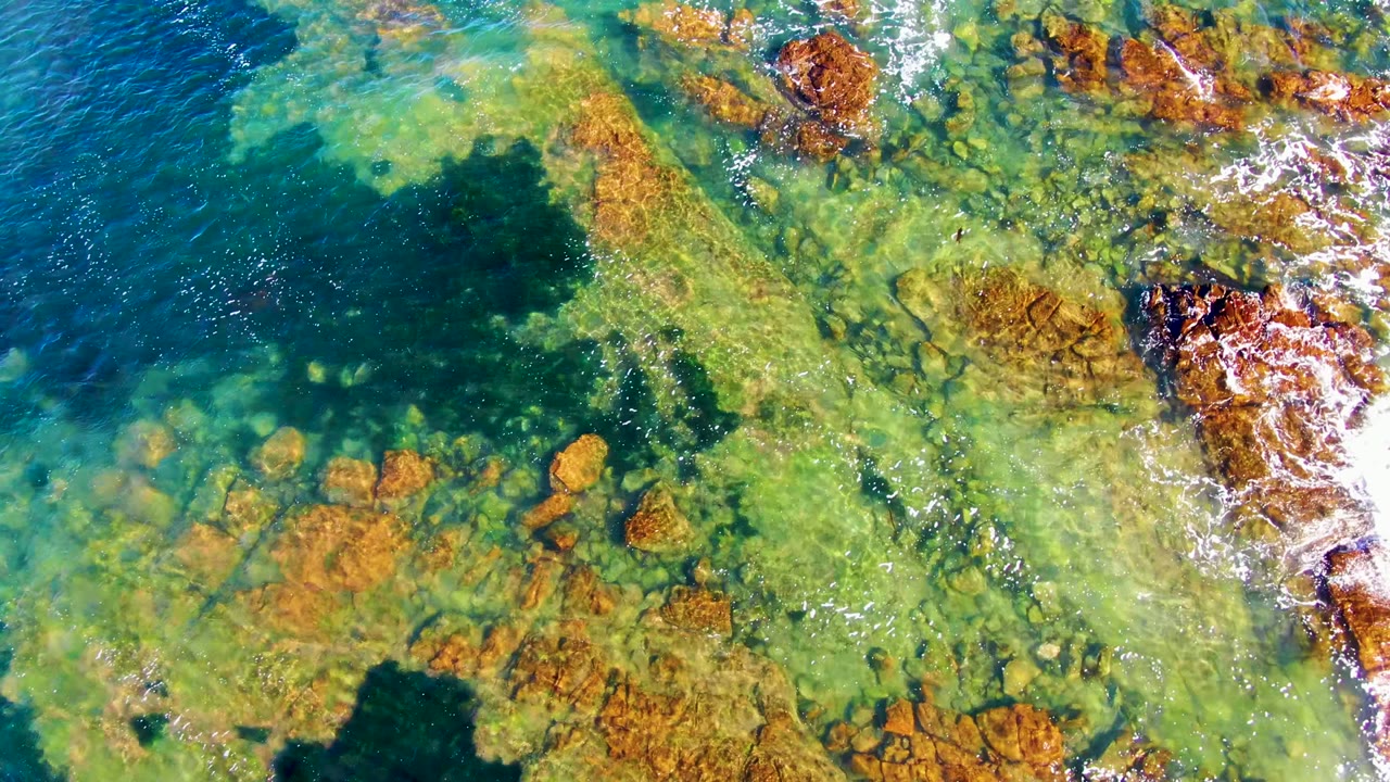 At 0:02, clear water in La Bocana exposes scattered rust-colored rocks while white wave foam brushes the rocky right edge, with no visible people.