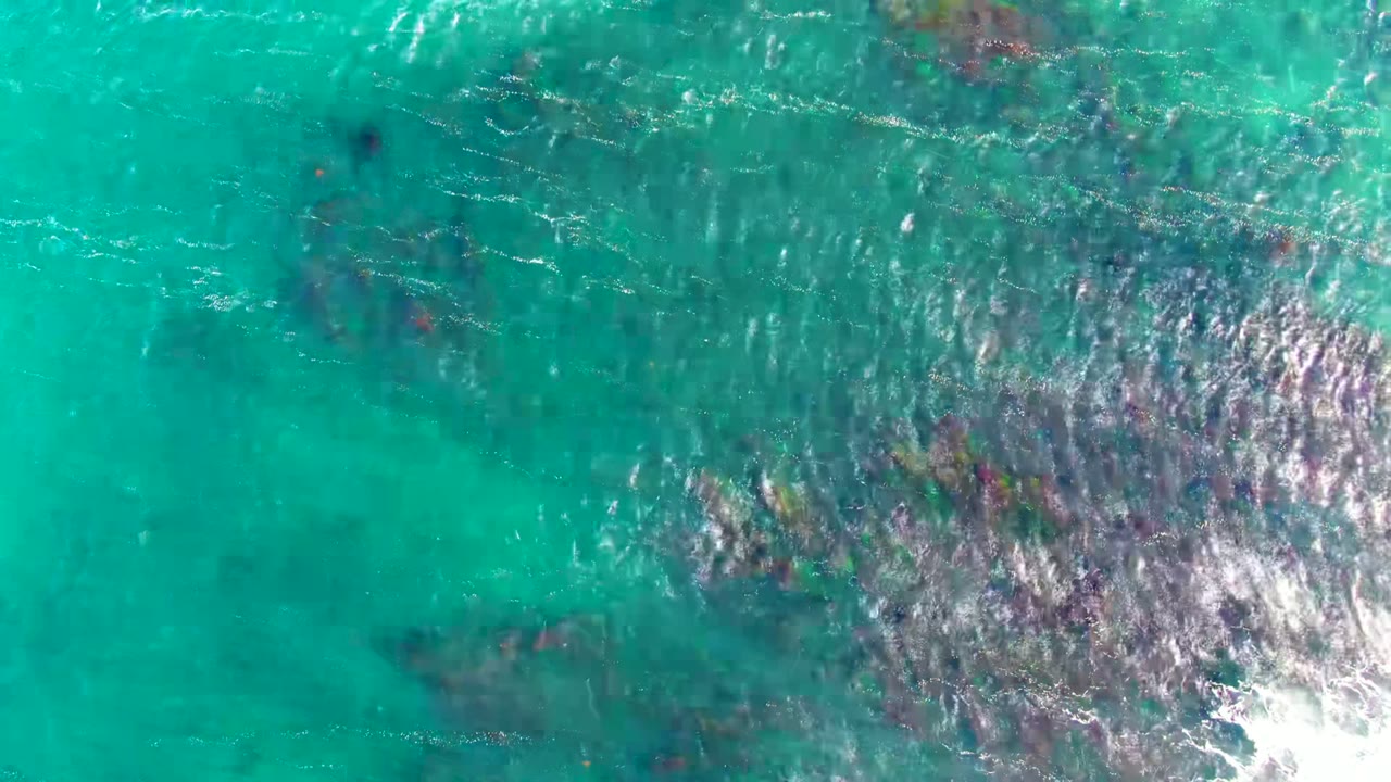 Overhead view of turquoise ocean water in La Bocana with dark rocky seabed patches and bright surface glare, no visible people.