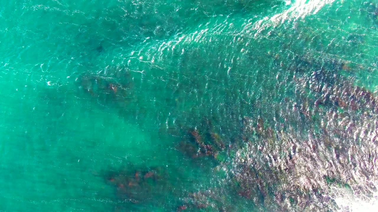 Overhead view of La Bocana water with dark reeflike seabed patches under turquoise water and white highlights crossing the upper right, no visible people.