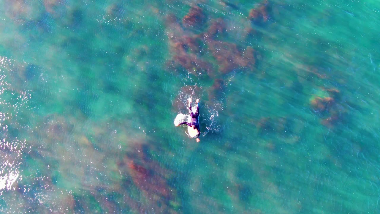 Overhead view of a swimmer in clear turquoise water at La Bocana, Mexico, with darker submerged rock or reef patches around the center of the frame.