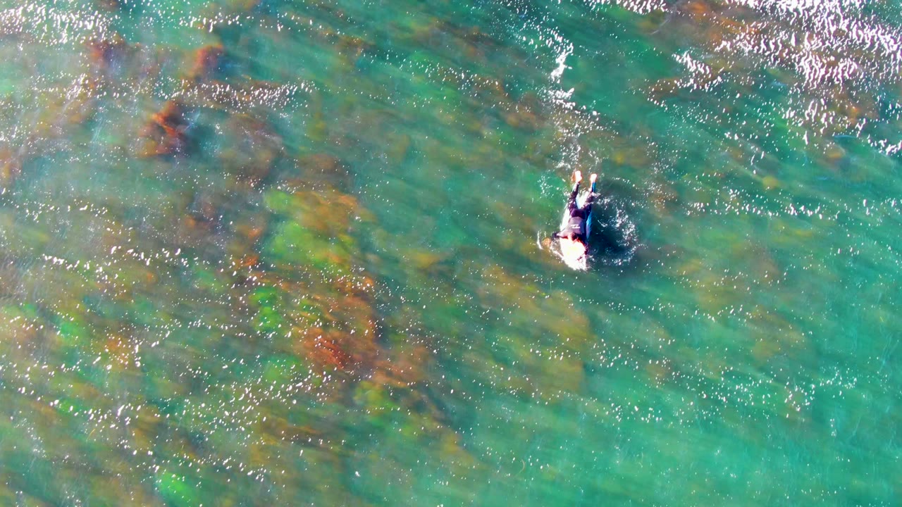 Top-down view of a swimmer in clear turquoise water at La Bocana, Mexico, with mottled brown and green underwater rock or coral patches beneath the surface.