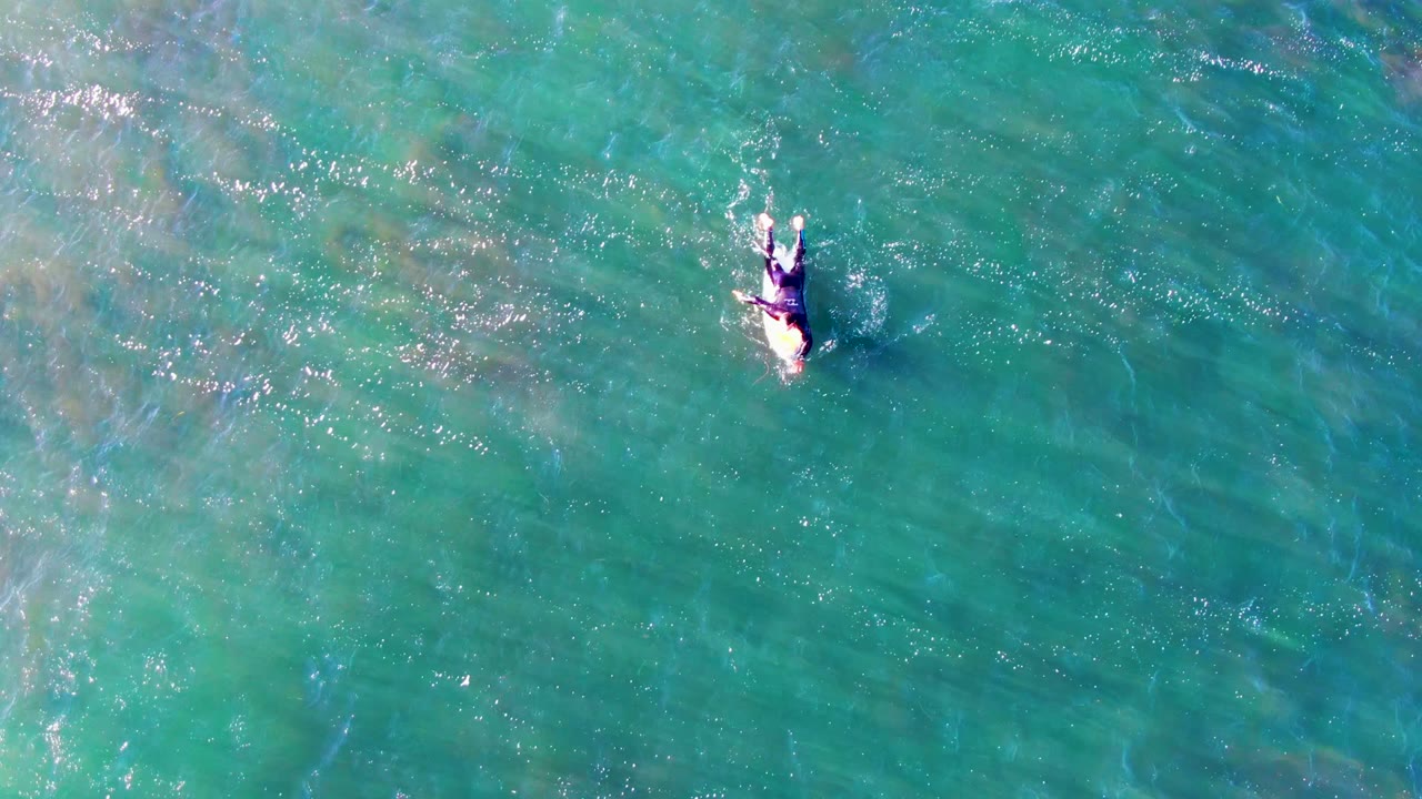 A swimmer seen from directly above in clear turquoise water at La Bocana, Mexico, with small white splashes around the body and faint seabed texture below.