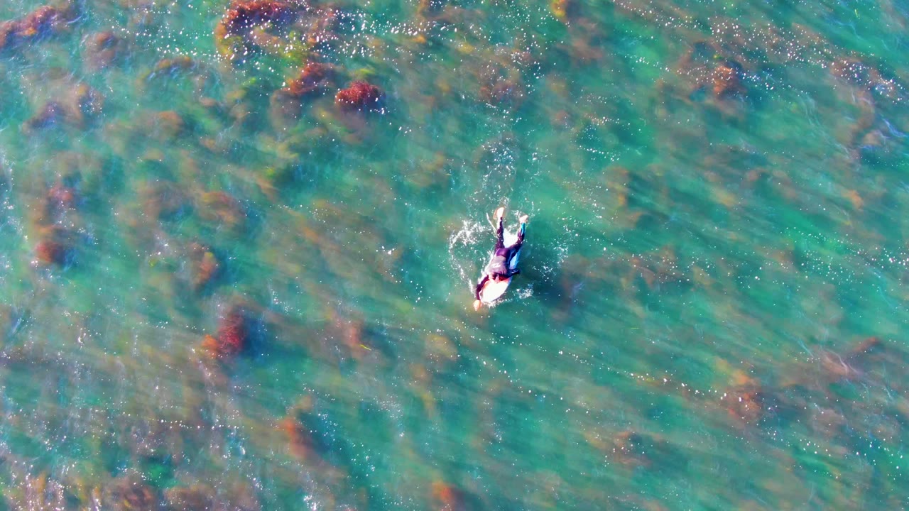 Overhead view in La Bocana, Mexico of one swimmer moving through turquoise shallow water with darker underwater rocks or coral visible below.