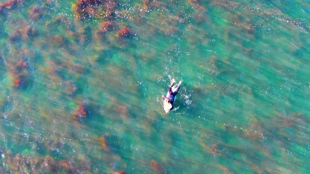 Aerial frame from La Bocana, Mexico showing the swimmer slightly farther across clear green-blue water, with mottled underwater rock or coral patches around them.