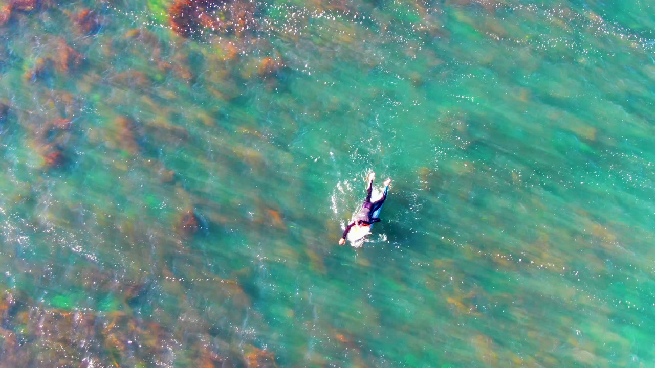 Top-down La Bocana, Mexico view of one swimmer in bright turquoise water, with reddish-brown and dark submerged rock or coral shapes visible beneath the surface.