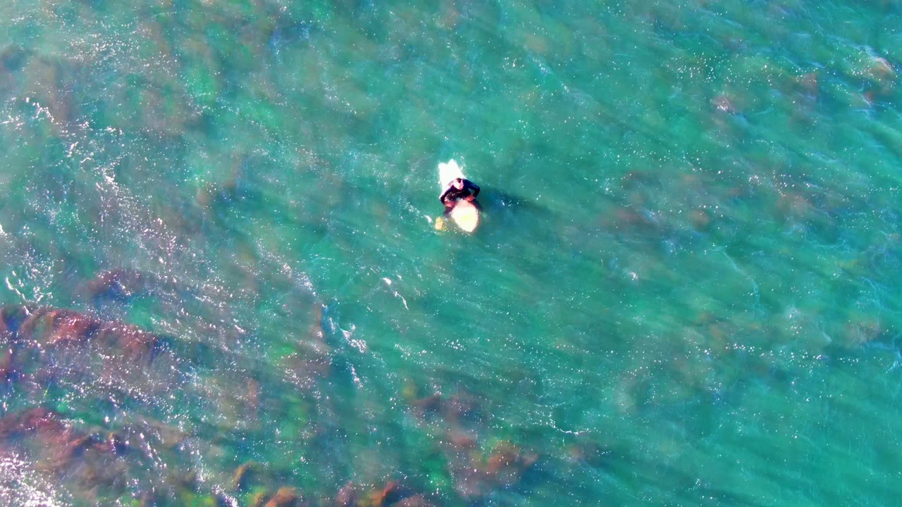 Overhead frame from La Bocana, Mexico, showing a person on a pale board near the upper center of clear turquoise water with darker seabed patches below.