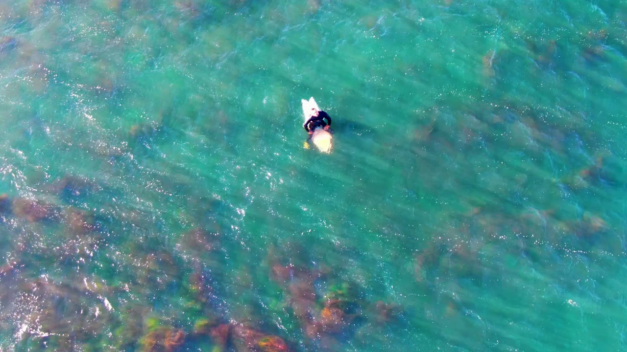 At two seconds, the overhead La Bocana view places the person and pale board above a wider spread of darker coral or rock patches beneath the turquoise surface.