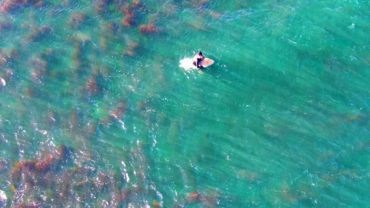 Overhead La Bocana, Mexico view with the person and white bodyboard near the upper right of the frame, surrounded by green-blue water and faint submerged kelp texture.