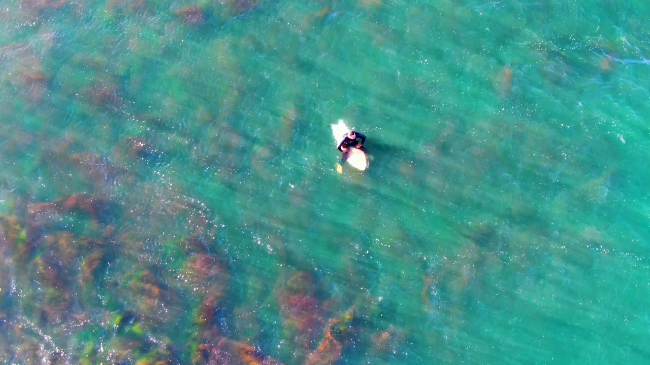 Overhead frame from La Bocana, Mexico showing a person on a light surfboard near the upper center, surrounded by turquoise water and darker underwater patches.