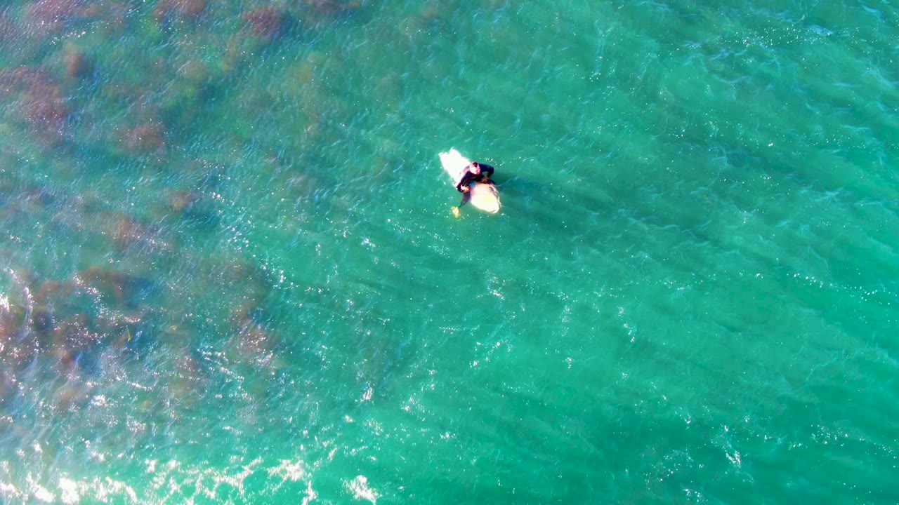 Overhead La Bocana frame with the person and surfboard near the upper middle right, with broad turquoise water and darker underwater areas spread across the left side.