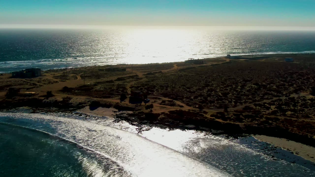 Opening aerial frame over La Bocana, Baja California Sur, with bright ocean glare, a sandy rocky shoreline, scrub-covered land, dirt paths, small structures, and no visible people.