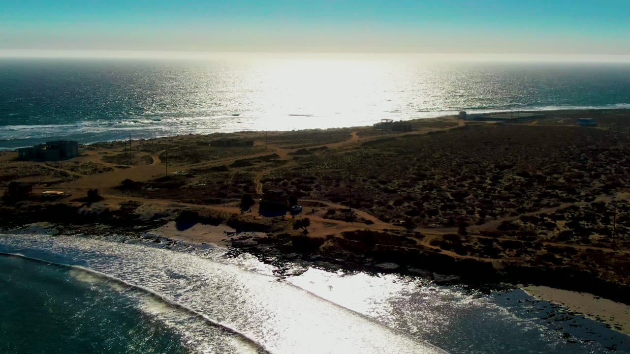 Backlit aerial frame over La Bocana, Baja California Sur, showing white surf near the beach, darker rocky shoreline, low vegetation, faint paths, small coastal structures, and no visible people.