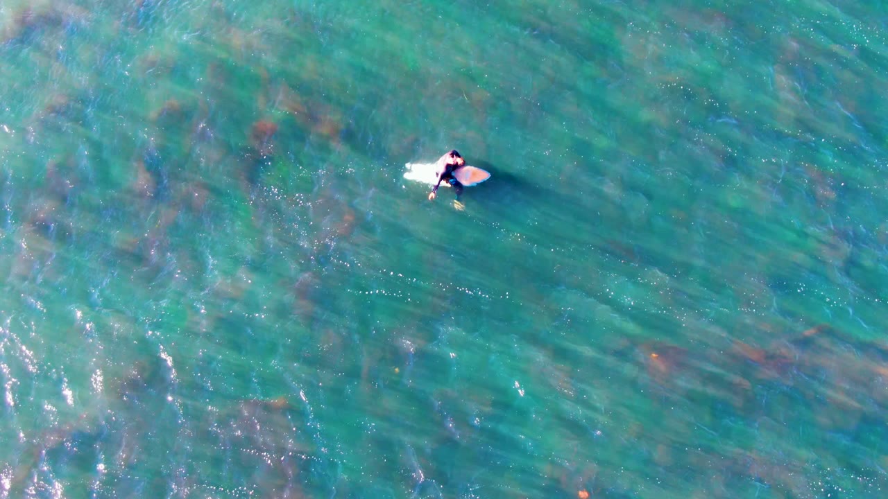 Overhead aerial frame from La Bocana, Mexico showing a person on a light surfboard near the upper left area of clear blue-green ocean water.