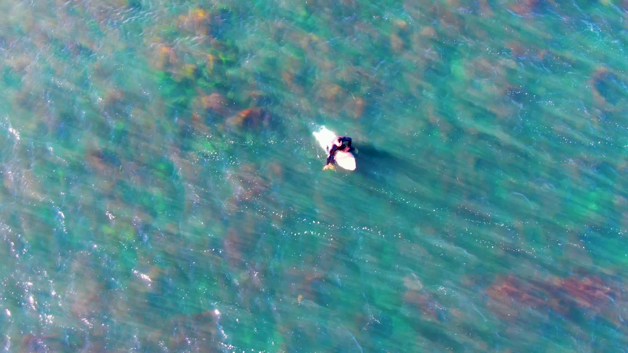Overhead La Bocana frame showing the person and surfboard near the upper right, with rippled turquoise water and darker underwater patches filling the frame.